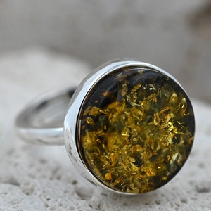 May include: A silver ring with a large, circular amber stone. The amber is a mix of golden and dark brown, with a textured, fragmented look. The ring's band and setting are made of polished silver.