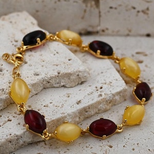 Könnte beinhalten: Ein goldenes Armband mit abwechselnd bernsteinfarbenen und roten ovalen Steinen.