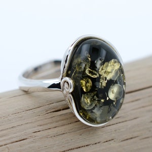 Può includere: Un anello d'argento con una pietra ovale di ambra verde. La pietra presenta un motivo a spirale di inclusiones nere e dorate.