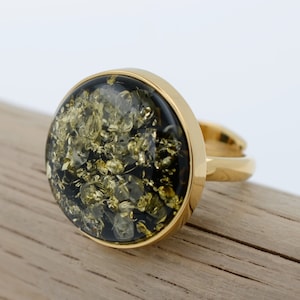 May include: A gold ring featuring a round, black and green stone set in the centre. The stone has a distinctive pattern of gold flecks throughout.