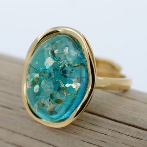 May include: A gold ring featuring a large oval turquoise blue stone set in a gold bezel. The stone has a sparkly, glittery effect.