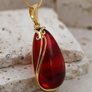 Op de afbeelding: Een druppelvormige hanger met een dieprode edelsteen, gevat in een goudkleurige zetting. De hanger hangt aan een fijne gouden ketting. De edelsteen heeft een gepolijste, doorschijnende uitstraling en de gouden zetting heeft decoratieve filigraan.