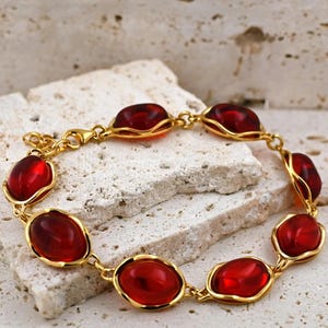 May include: A gold bracelet featuring red oval-shaped glass stones. The bracelet has a lobster claw clasp.