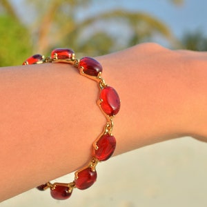 Op de afbeelding: Een gouden armband met rode ovale stenen. De armband zit om de pols van een persoon.