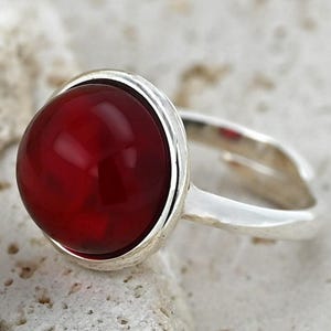 May include: A silver ring featuring a large, round, deep red gemstone. The gemstone is set in a bezel setting, and the ring band is a simple, polished silver. The ring is displayed on a textured, light-colored surface.