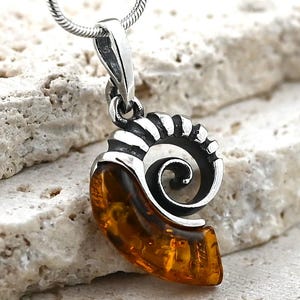 May include: A silver pendant necklace with an ammonite fossil design. The pendant has a silver frame and a central section of amber. The necklace chain is a thin, silver snake chain. The amber is a warm, translucent colour.