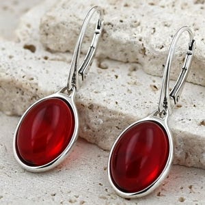 May include: Silver leverback earrings featuring oval, deep red cabochon stones. The earrings are set against a textured, light-coloured stone surface. The design is simple and elegant, suitable for everyday wear or special occasions.