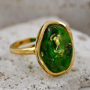 May include: A gold ring with a large, oval green stone. The stone has a bubbly texture and is set in a gold bezel. The ring's band is plain, polished gold. A piece of jewellery.