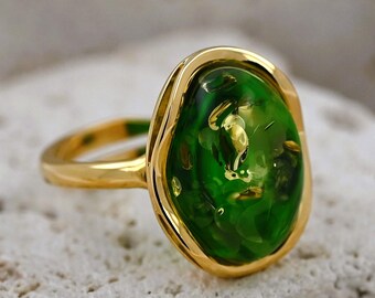 May include: A gold ring featuring a large, oval-shaped green stone. The stone has a bubbly, textured appearance and is set within a gold bezel. The ring's band is a simple, polished gold.