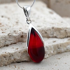 May include: A teardrop-shaped pendant with a vibrant red center, encased in a polished silver frame. The pendant hangs from a delicate silver chain. The red color transitions from a lighter shade at the top to a deeper hue at the bottom, creating a striking visual effect.