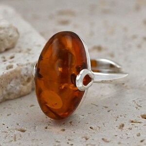 May include: A silver ring with a large, oval amber gemstone. The amber is a warm, translucent orange-brown with inclusions. The ring has a simple design with a split shank and decorative accents. The amber measures approximately 2.5 cm in length.