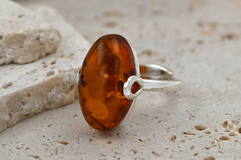 May include: A silver ring with a large, oval amber gemstone. The amber is a warm, translucent orange-brown with inclusions. The ring has a simple design with a split shank and decorative accents. The amber measures approximately 2.5 cm in length.
