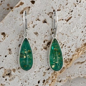 May include: A pair of silver dangle earrings with green teardrop-shaped resin stones. The stones have a sparkly gold fleck throughout.