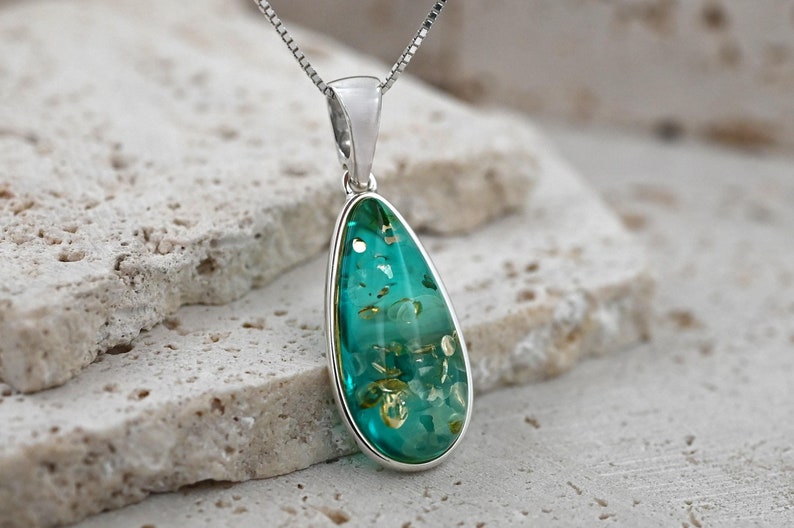 May include: A teardrop-shaped pendant with a silver setting and chain. The pendant features a translucent teal stone with embedded golden flecks. The jewelry is displayed on a textured, light-colored surface.