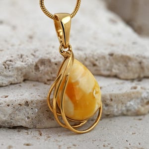 May include: A gold pendant necklace with a teardrop-shaped amber stone. The amber has a creamy white and orange hue. The pendant hangs from a fine gold chain. The pendant is set within a gold metal frame.