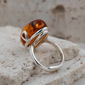 May include: A silver ring with a large, oval amber gemstone. The amber is a warm, translucent orange-brown hue. The ring's band is smooth and polished, with a decorative silver setting around the amber. A piece of jewellery.
