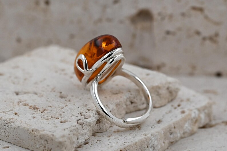 May include: A silver ring with a large, oval amber gemstone. The amber is a warm, translucent orange-brown hue. The ring's band is smooth and polished, with a decorative silver setting around the amber. A piece of jewellery.