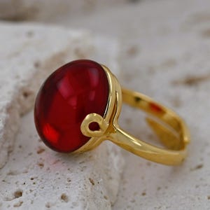 Könnte beinhalten: Ein goldener Ring mit einem großen, runden, roten Edelstein. Der Ring hat ein einzigartiges Design mit einem gebogenen Band und einem kleinen herzförmigen Detail.