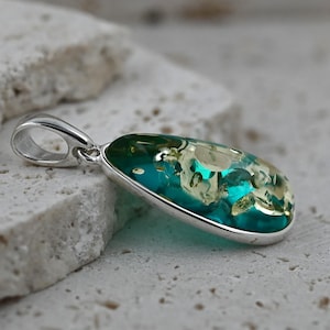 May include: A silver pendant with a teardrop-shaped, teal-colored stone. The stone has a mix of light green and white inclusions, and is set in a polished silver bezel. The pendant has a simple, elegant design.