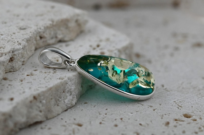 May include: A silver pendant with a teardrop-shaped, teal-colored stone. The stone has a mix of light green and white inclusions, and is set in a polished silver bezel. The pendant has a simple, elegant design.