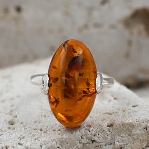 May include: An oval amber ring with a silver band. The amber stone is a warm, translucent orange colour with dark inclusions. The ring is set on a textured, light-coloured surface.