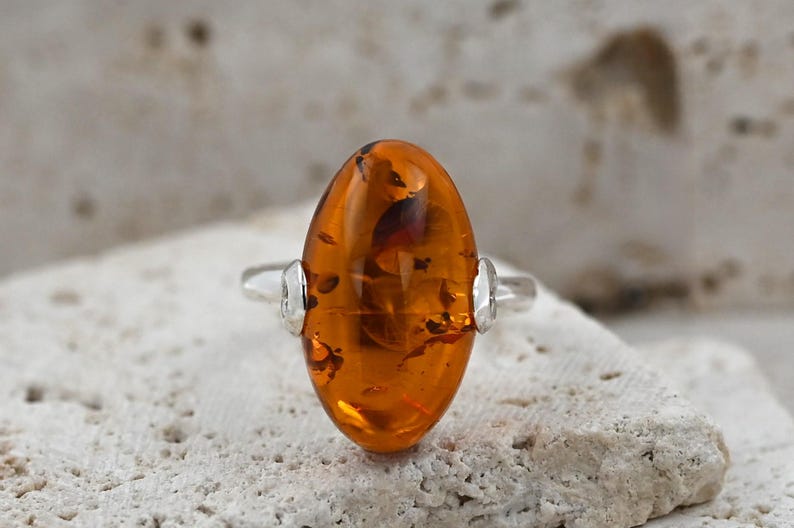 May include: An oval amber ring with a silver band. The amber stone is a warm, translucent orange colour with dark inclusions. The ring is set on a textured, light-coloured surface.