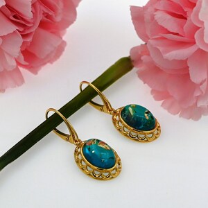 May include: A pair of gold earrings with blue and green oval-shaped stones set in a decorative gold frame. The earrings are hanging from a green stem with pink flowers.