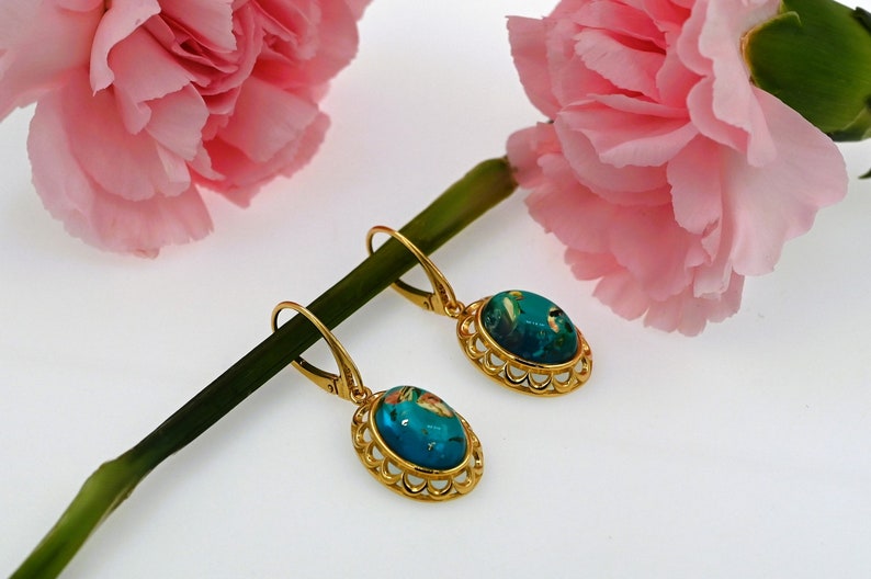May include: A pair of gold earrings with blue and green oval-shaped stones set in a decorative gold frame. The earrings are hanging from a green stem with pink flowers.