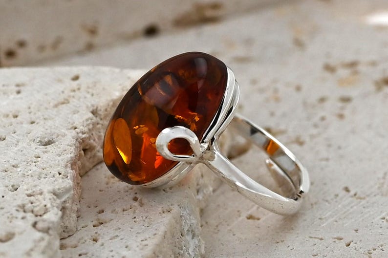 May include: A silver ring with a large, oval amber gemstone. The amber is a warm, translucent brown and orange colour. The ring has a simple, polished silver band and a decorative loop detail.