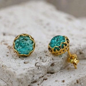 May include: A pair of gold-colored stud earrings with round, turquoise-colored stones. The earrings have a decorative, scalloped gold setting. The earrings are displayed on a textured, off-white surface.