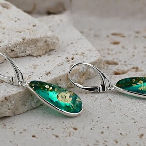May include: A pair of silver dangle earrings with teardrop-shaped green resin stones. The stones have a gold fleck pattern.