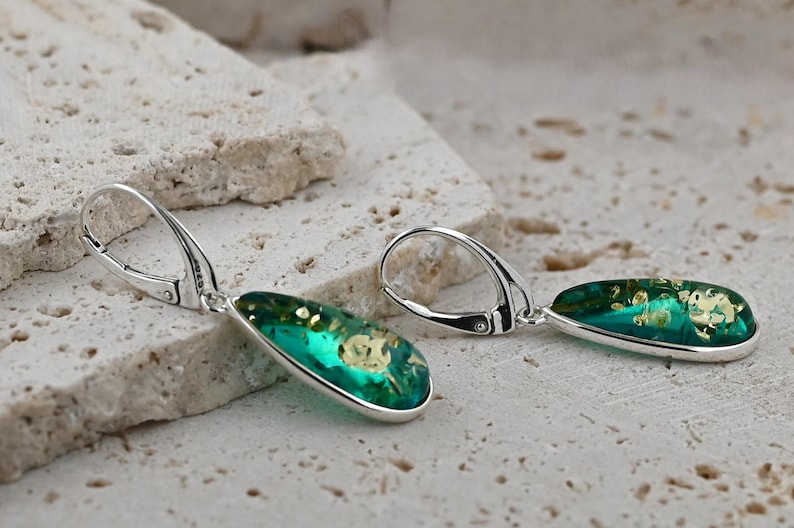 May include: A pair of silver dangle earrings with teardrop-shaped green resin stones. The stones have a gold fleck pattern.