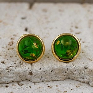 May include: A pair of round stud earrings. Each earring features a vibrant green, translucent orb encased in a gold-colored metal setting. The green orbs contain small, gold-colored flecks, adding a touch of sparkle.