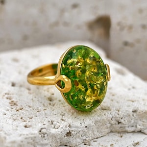 May include: A gold ring featuring an oval, green gemstone with gold inclusions. The ring's band and setting are gold. The ring is displayed on a white, textured surface. The gemstone is approximately 2 cm in length.