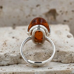 May include: A silver ring with a large, oval amber gemstone. The amber is a warm, translucent orange-brown colour. The ring's band is polished silver. The ring is set on a textured, light-coloured surface.