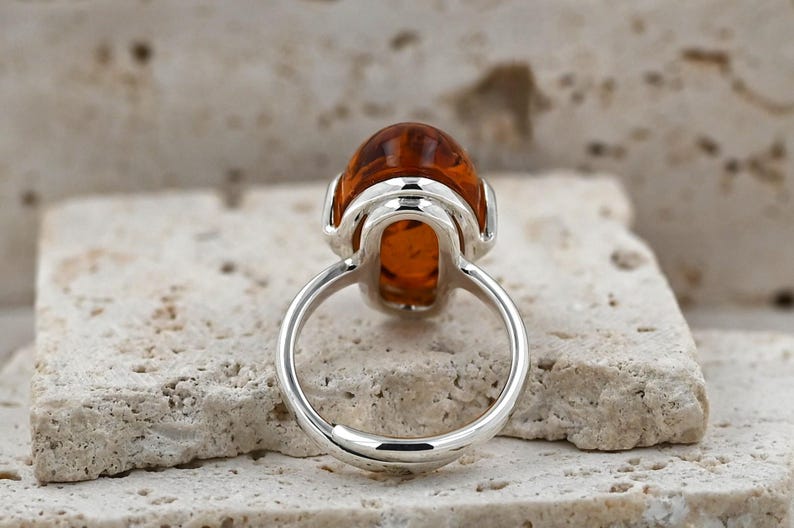 May include: A silver ring with a large, oval amber gemstone. The amber is a warm, translucent orange-brown colour. The ring's band is polished silver. The ring is set on a textured, light-coloured surface.