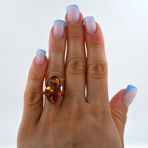 May include: A gold ring with a large, oval amber gemstone. The amber is a warm, honey-brown colour with internal patterns. The ring is on a hand with manicured nails, painted with a light pink and blue French manicure.