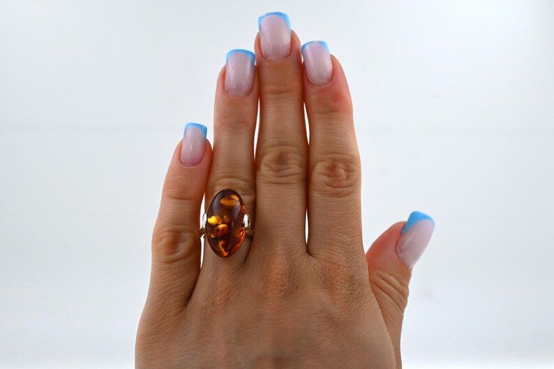 May include: A gold ring with a large, oval amber gemstone. The amber is a warm, honey-brown colour with internal patterns. The ring is on a hand with manicured nails, painted with a light pink and blue French manicure.