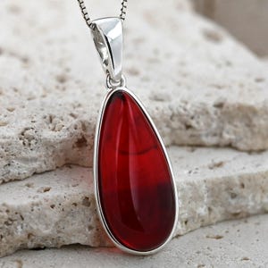 May include: A teardrop-shaped pendant with a vibrant red gemstone, encased in a silver setting. The pendant hangs from a delicate silver chain. The gemstone's color deepens towards the bottom, creating a striking visual contrast.