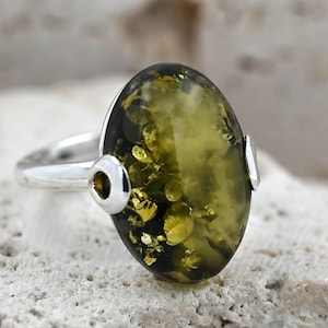 May include: A sterling silver ring featuring an oval-shaped amber stone. The amber has a dark green base with golden inclusions. The ring's band is smooth, and it has decorative silver accents on either side of the amber stone.