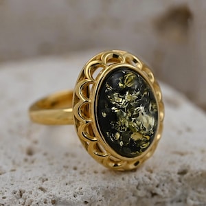 May include: A gold ring with an oval, dark green amber stone with golden inclusions. The ring has an openwork setting and a polished gold band. The ring is a piece of jewellery.