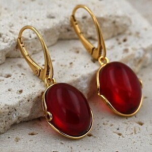 May include: A pair of gold-colored leverback earrings with oval, deep red gemstones. The gemstones are set in a gold-colored bezel. The earrings are displayed on a textured, light-colored surface.