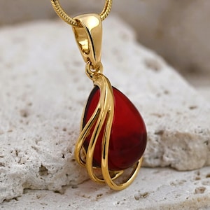 May include: A gold pendant necklace with a teardrop-shaped, deep red gemstone. The gemstone is held in a gold, open-work setting, hanging from a fine gold chain. The pendant is shown against a textured, light-coloured surface.