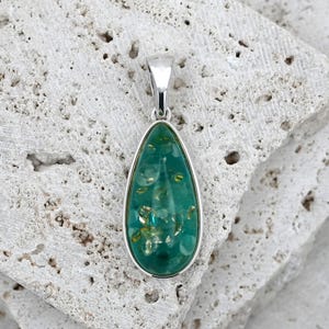 May include: A teardrop-shaped pendant with a silver-colored bail and frame. The pendant's center is a vibrant teal color with embedded gold-colored flecks. The pendant is resting on a textured, light-colored stone surface.