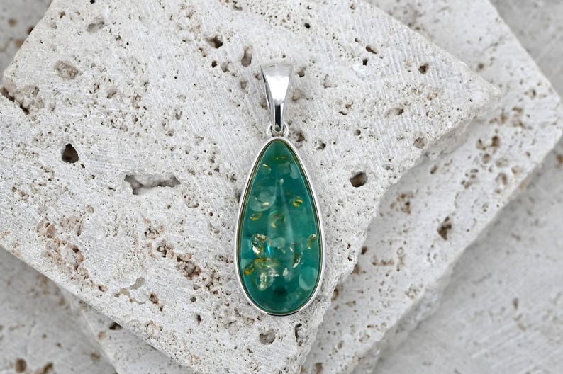 May include: A teardrop-shaped pendant with a silver-colored bail and frame. The pendant's center is a vibrant teal color with embedded gold-colored flecks. The pendant is resting on a textured, light-colored stone surface.