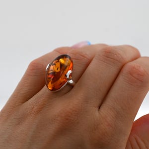 May include: A silver ring with a large, oval amber gemstone. The amber has a warm, golden-orange colour with internal patterns. The ring is set on a hand with manicured nails, against a white background.