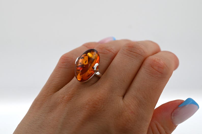May include: A silver ring with a large, oval amber gemstone. The amber has a warm, golden-orange colour with internal patterns. The ring is set on a hand with manicured nails, against a white background.