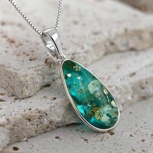 May include: A teardrop-shaped pendant necklace with a silver chain. The pendant has a silver bezel and contains a teal-colored, translucent material with embedded golden and white inclusions. The necklace rests on a light-colored, textured surface.