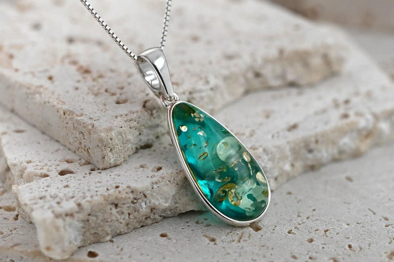 May include: A teardrop-shaped pendant necklace with a silver chain. The pendant has a silver bezel and contains a teal-colored, translucent material with embedded golden and white inclusions. The necklace rests on a light-colored, textured surface.