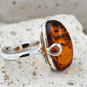 May include: Silver ring with a large, oval amber gemstone. The amber is a warm, translucent orange-brown hue. The ring has a simple, elegant design with a polished silver band and decorative detailing around the stone.